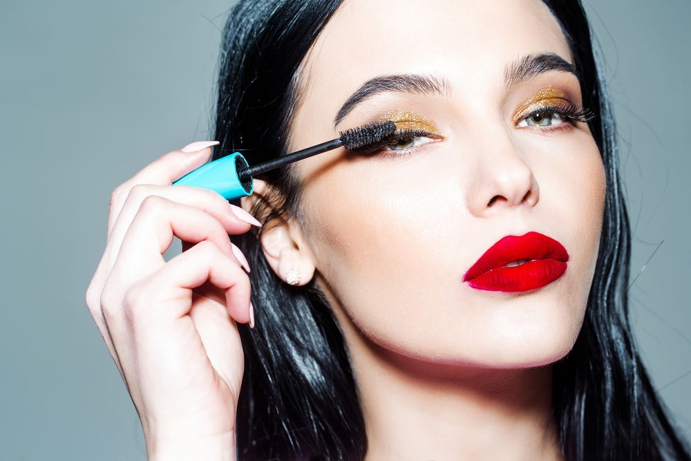 Pale woman with dark hair and red lipstick applying mascara.